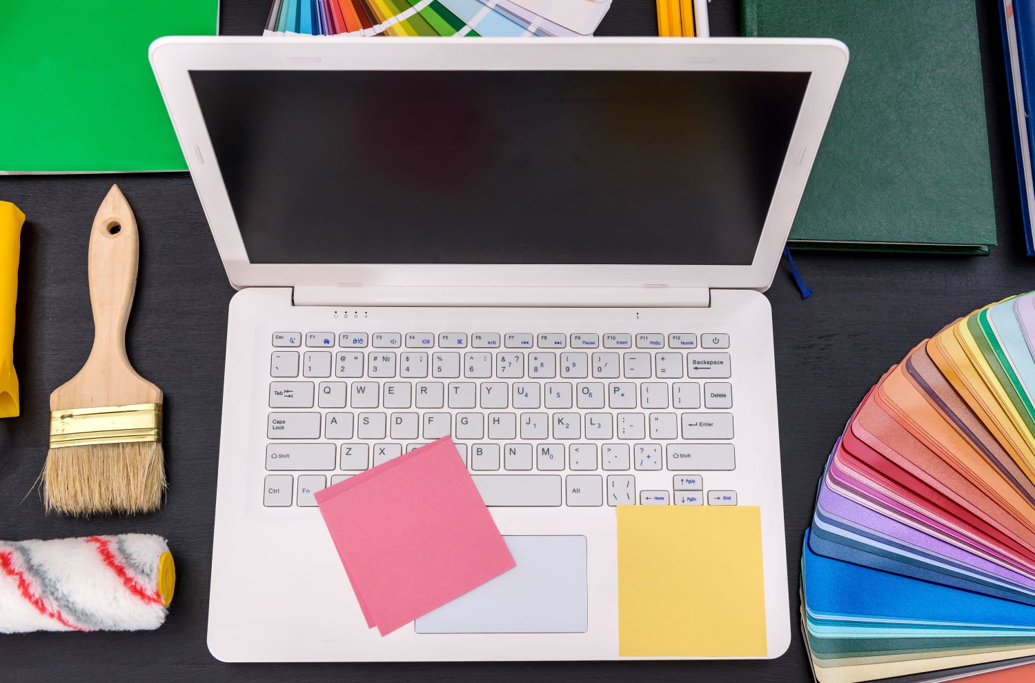 laptop open with a paint brush, paint roller and colour paint swatch cards next to the laptop